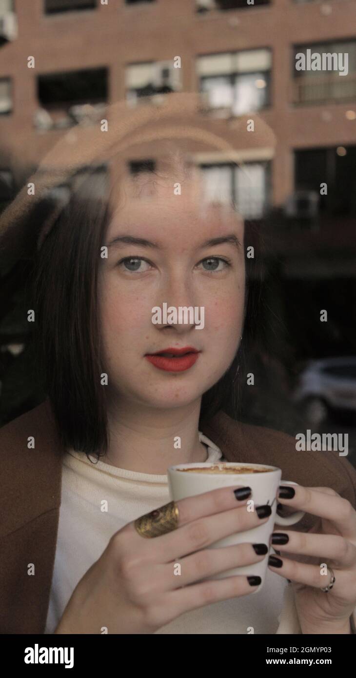 Giovane donna in un bar che tiene in mano una tazza calda di caffè pronto a bere in posa per la macchina fotografica. Stile Vibes parigino per la stagione autunnale 2021. Foto Stock