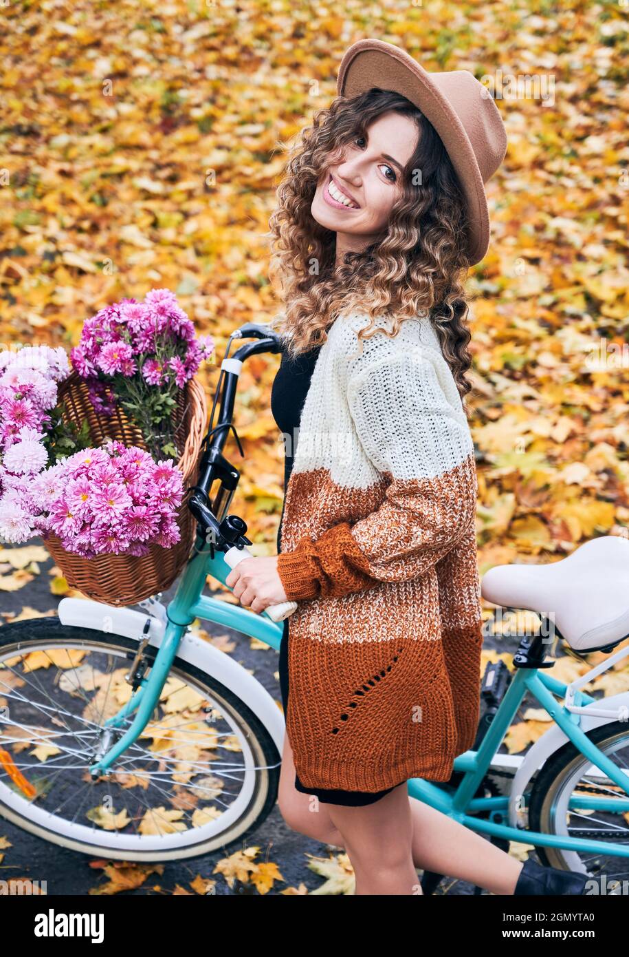 Vista laterale della bruna curly viaggiando in bicicletta d'epoca con un grande cesto di fiori rosa. Bella donna che indossa cappello marrone e cardigan è in bicicletta nel parco autunnale. Concetto di fine settimana . Foto Stock