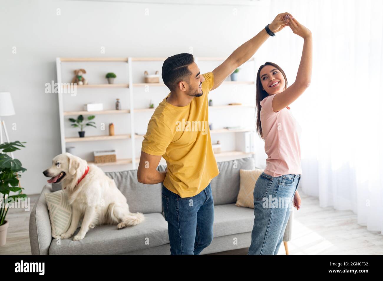 Romantico giovane coppia diverse ballare a casa, il loro cane carino seduto sul divano vicino, spazio copia Foto Stock
