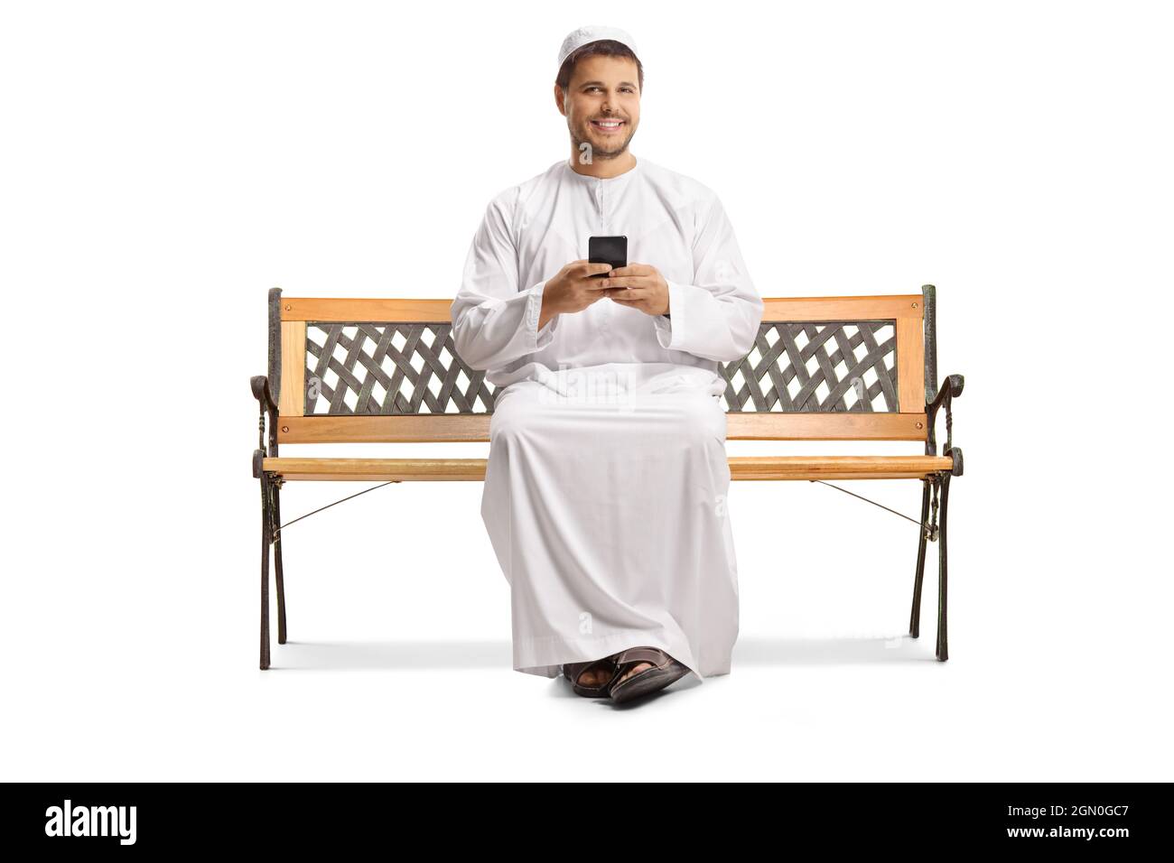 Uomo in abiti etnici seduto su una panca con uno smartphone e guardando la macchina fotografica isolato su sfondo bianco Foto Stock