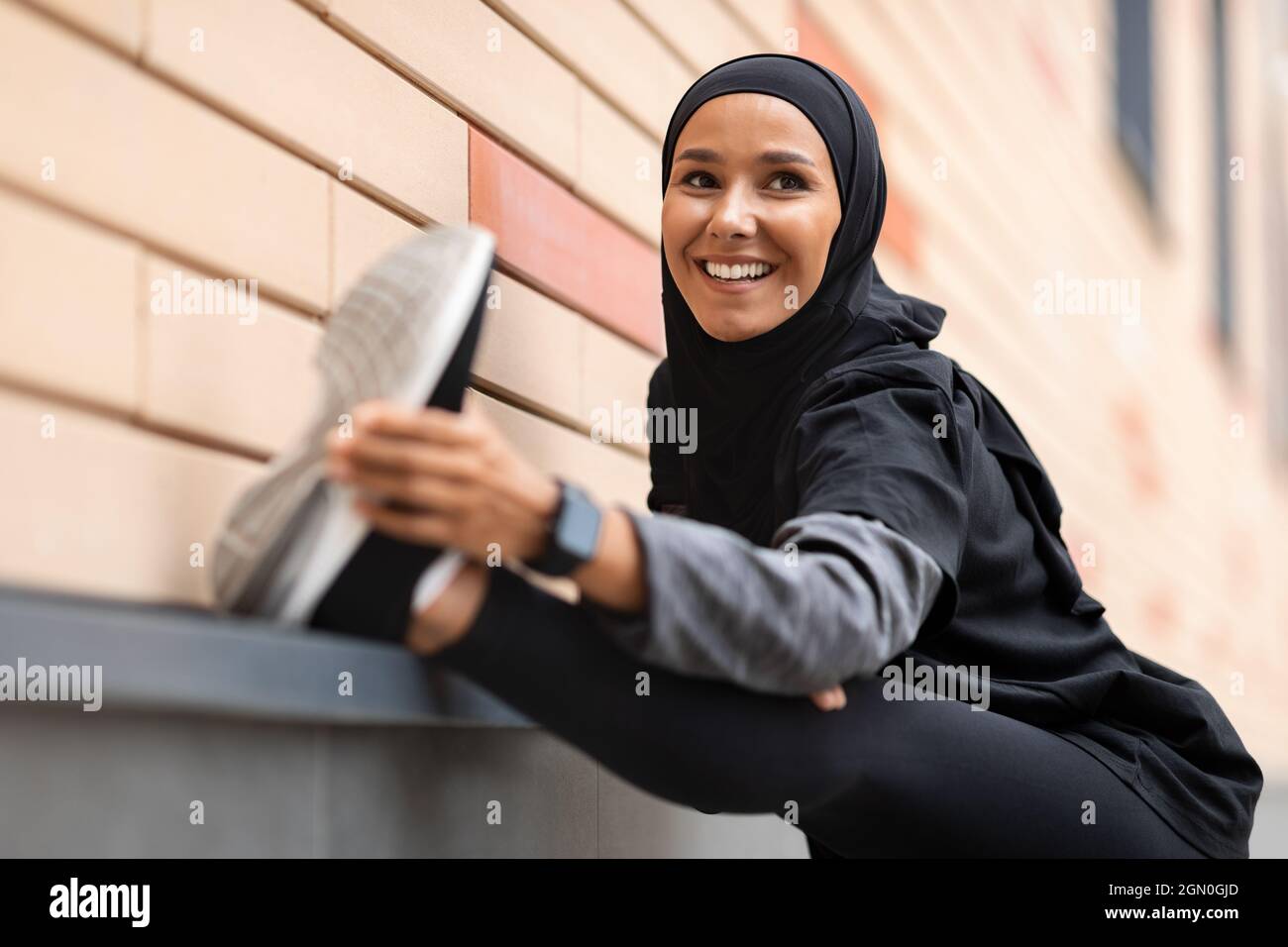 Donna musulmana sorridente e sportiva in Hijab che allunga i muscoli delle gambe prima di allenarsi all'aperto Foto Stock