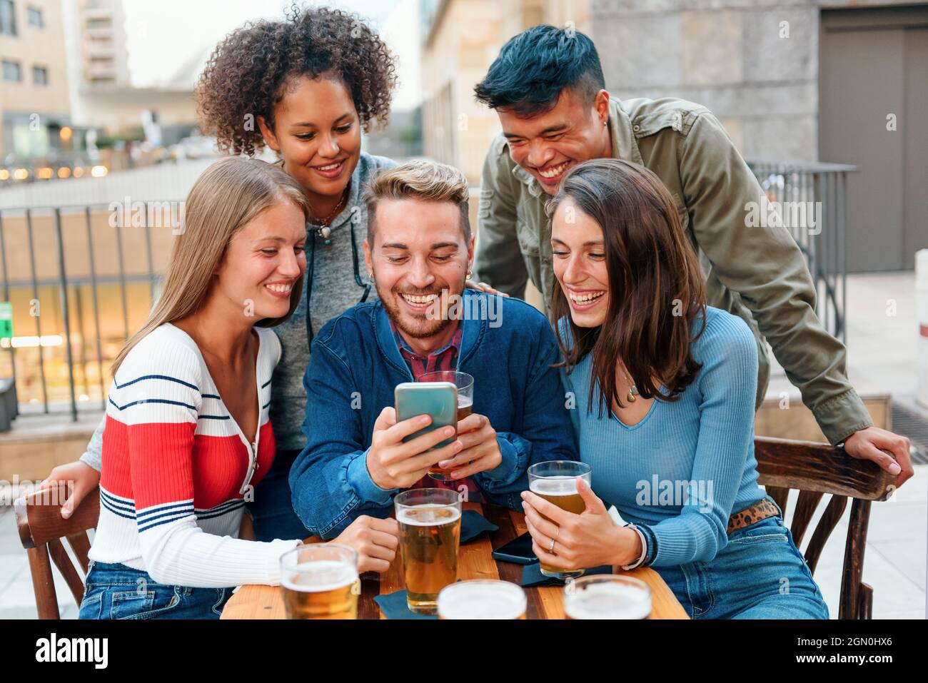Un gruppo di giovani amici diversi si è riunito intorno a uno smartphone a un tavolo da pub mentre si godono una birra insieme ridendo e scherzando mentre fanno una videochiamata Foto Stock