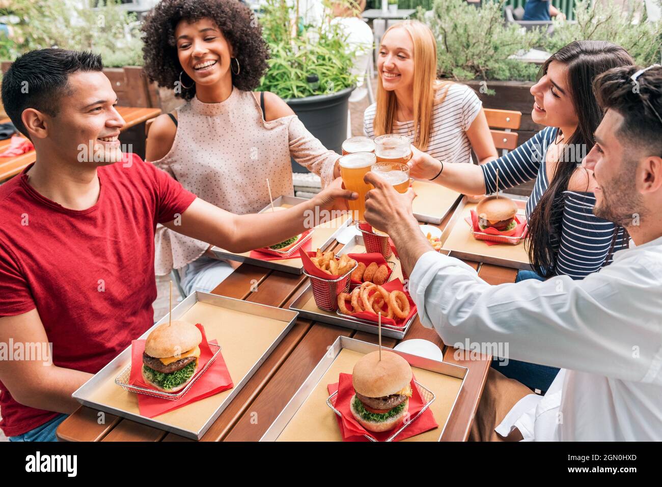 Gruppo di amici allegri multietnici che si riuniscono a tavola con fast food e bicchieri di birra aggraffati, mentre propongono pane tostato e festeggia un amico Foto Stock