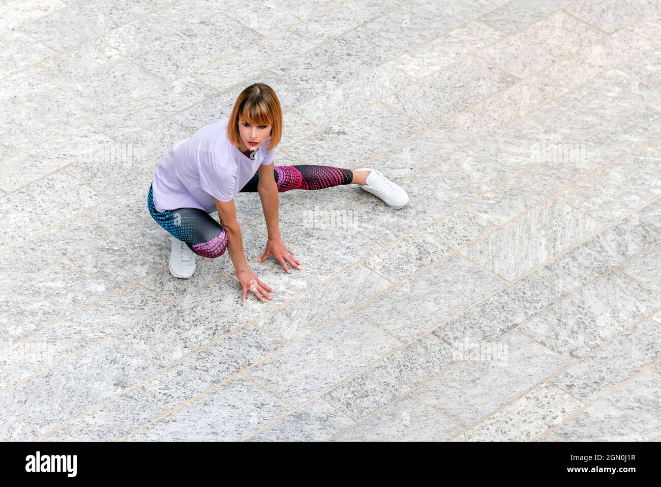 Da sopra tutto il corpo di giovane donna vestita in elegante abbigliamento sportivo e sneaker che fanno esercizio stretching per le gambe su quadrato urbano pavimentato durante la fi all'aperto Foto Stock