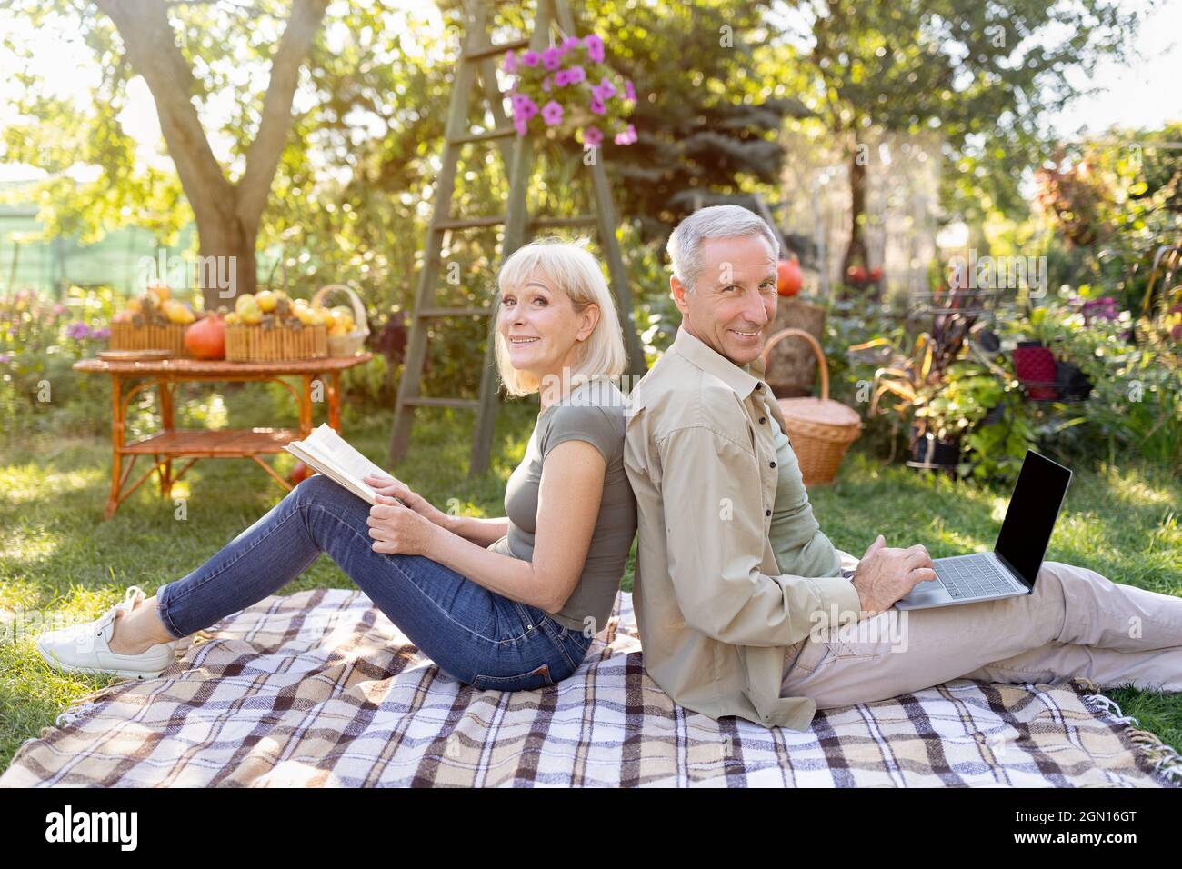 Coppia anziana felice che riposa su coperta da picnic in giardino, uomo che usa il computer portatile, donna che legge libro Foto Stock