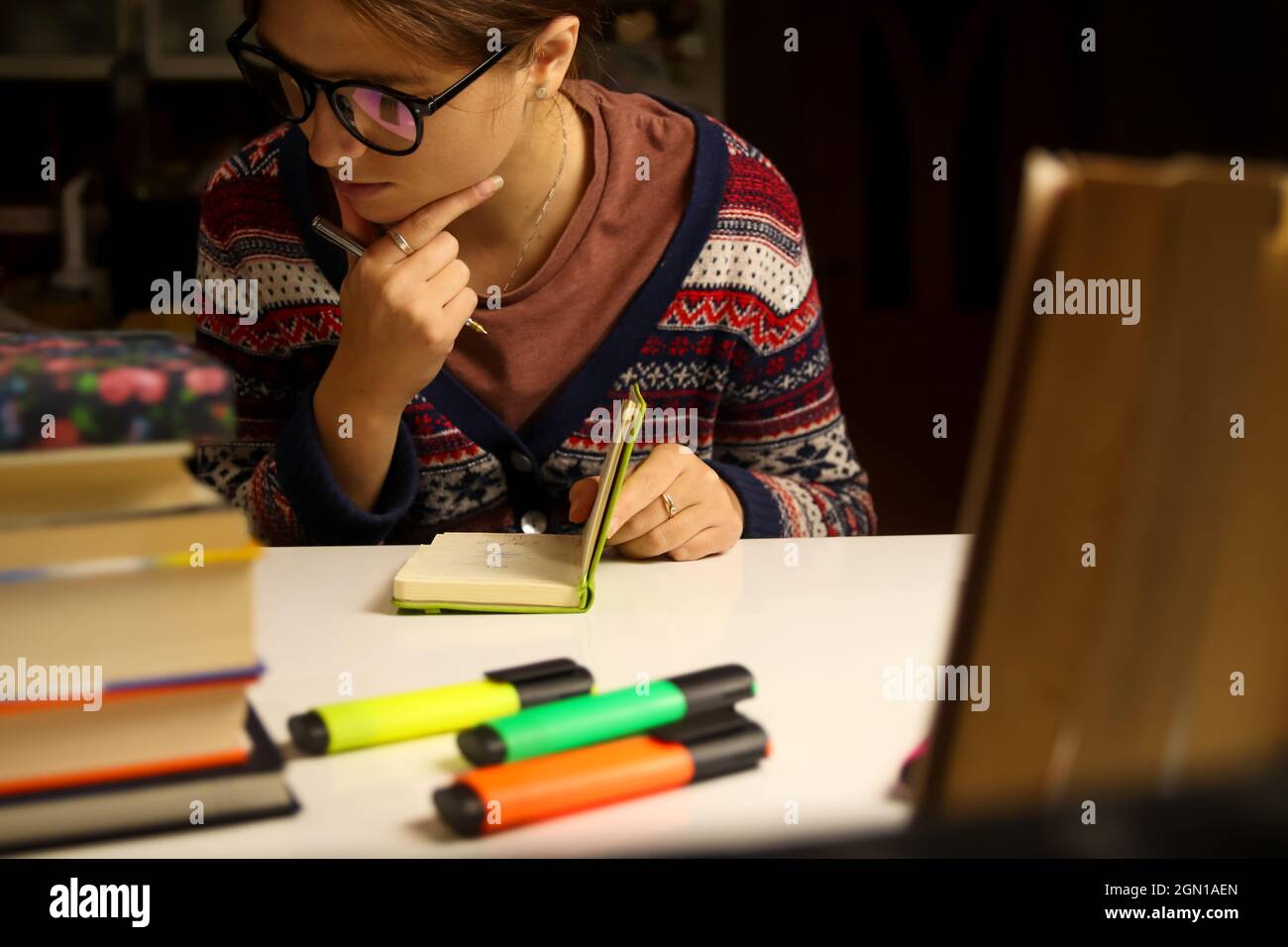 Giovane donna in occhiali studio di notte, pensa. Studente millenario con notebook verde chiaro. Libri e marcatori sfocati in primo piano. Istruzione e. Foto Stock