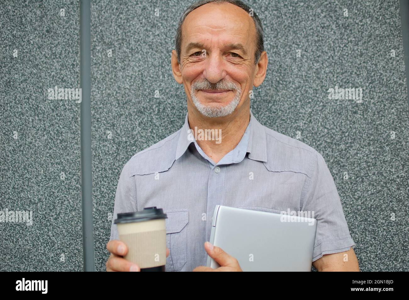 Ritratto maschile del collega più anziano con computer portatile argento e tazza di caffè in attesa di una riunione vicino al muro grigio scuro al mattino, lavoro remoto, caffè Foto Stock