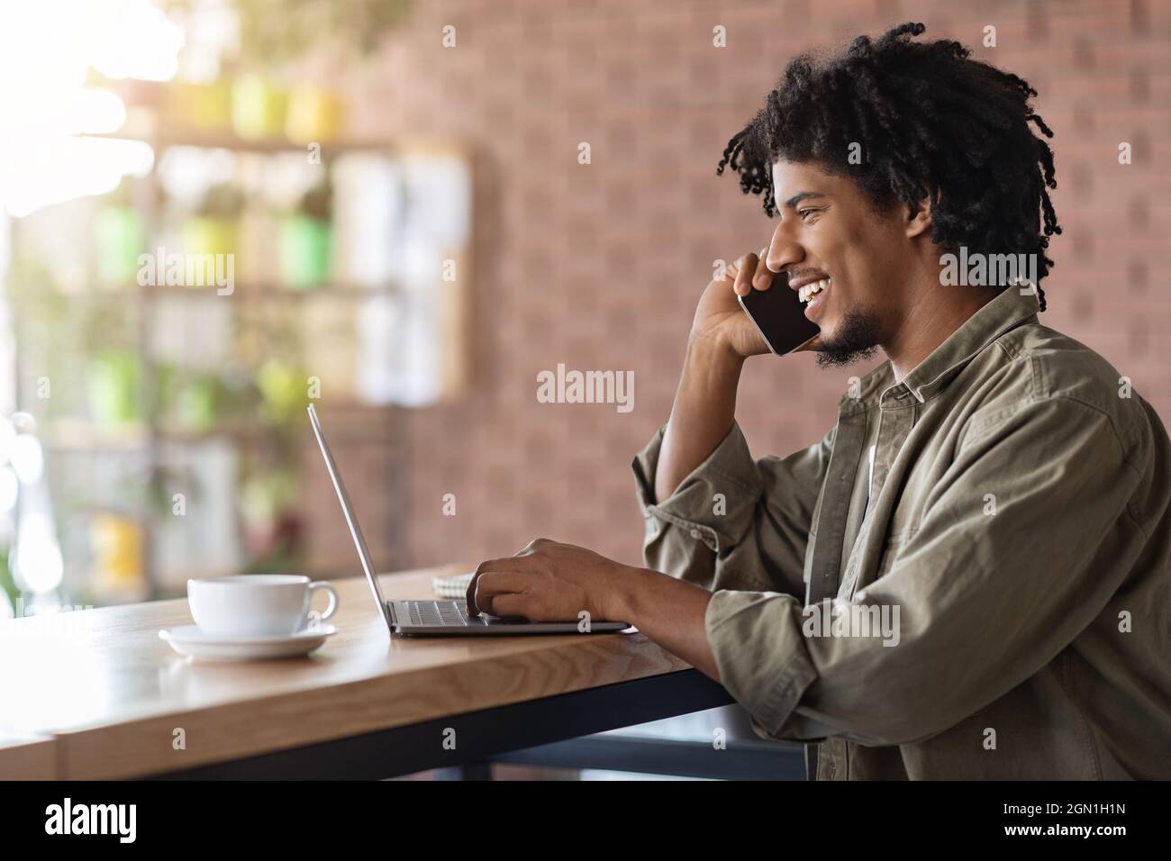 Black Freelancer Guy parla con il cellulare mentre lavora con il laptop al Cafe Foto Stock