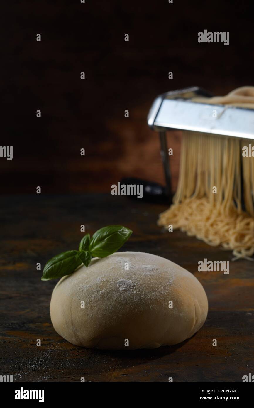 Pasta cruda con foglie di basilico posta su tavola di legno contro macchina metallica per la produzione di spaghetti in studio scuro Foto Stock