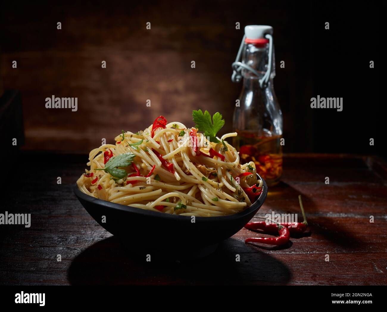 Deliziosi spaghetti aglio e olio con peperoncino piccante e prezzemolo serviti in ciotola su tavola di legno in studio scuro Foto Stock