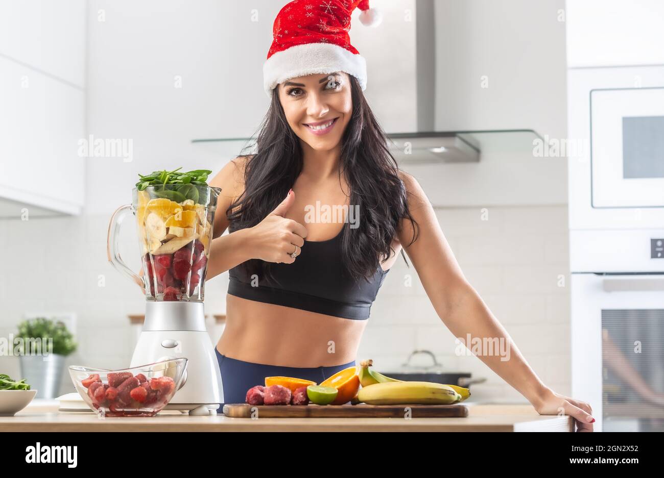 Pollice su da una donna in cappello di Babbo Natale in piedi in abiti da allenamento accanto a sano smoothie maker pieno di roba sano. Foto Stock