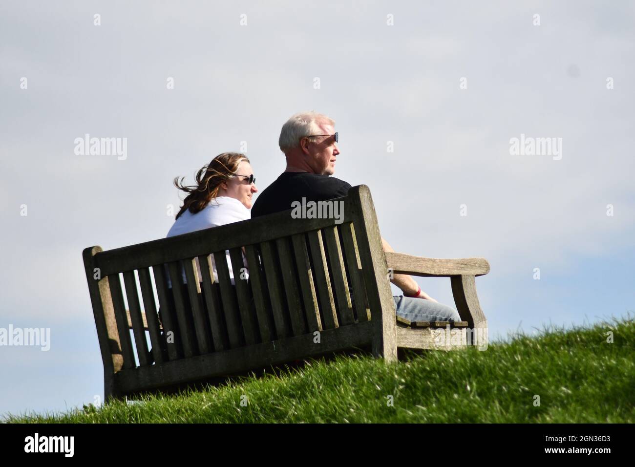 Una coppia matura seduta su una panca di legno sulla cima di una banca di erba, entrambi guardando a destra con il vento che soffia in giornata nuvolosa di sole Foto Stock