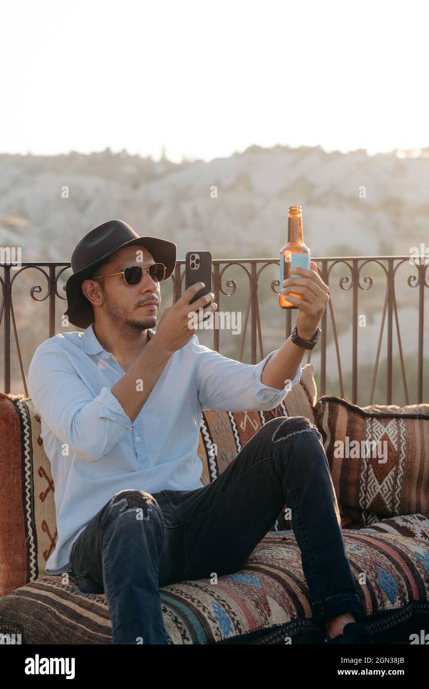 Uomo moderno in elegante vestito e occhiali da sole seduti su cuscini e fotografare una bottiglia di birra di vetro sul cellulare sul bar terrazza in Cappadocia, Foto Stock