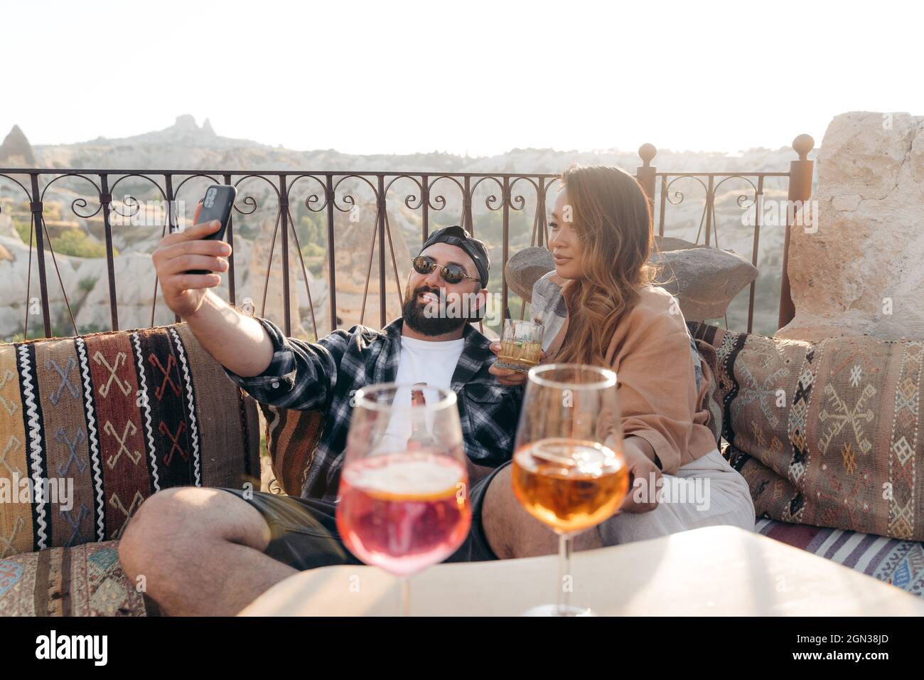 Positivo giovane coppia elegante seduta sul divano e bicchieri di cocktail reclinabili mentre si prende autoritratto sul cellulare in terrazza in Cappadocia, Turk Foto Stock