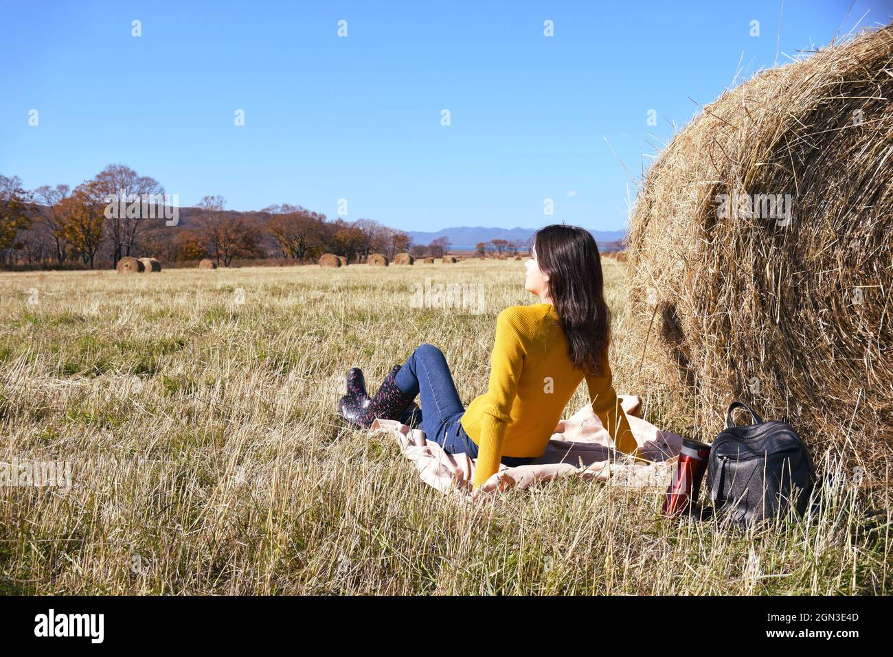 Donna in maglione giallo e stivali di gomma seduta su plaid accanto a zaino e thermos con tè caldo vicino haystack in campo Foto Stock