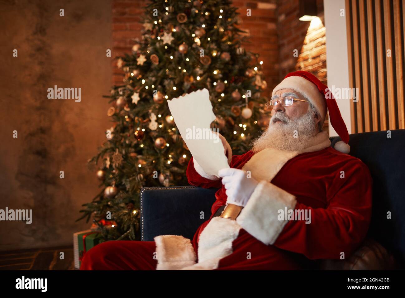 Squinting Babbo Natale in occhiali seduti sul divano a illuminato albero di Natale e lettura vecchia lettera Foto Stock