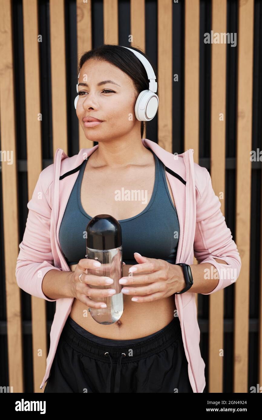 Bellissima donna africana in cuffie wireless che tiene una bottiglia d'acqua durante l'allenamento all'aperto Foto Stock