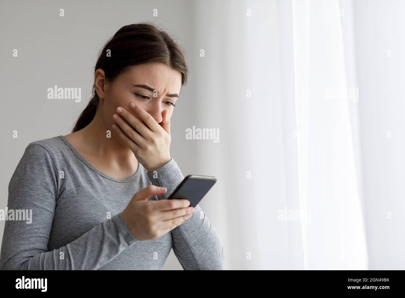 Sconvolto la donna graziosa millenaria depressa che copre la sua bocca con la mano e guardando il telefono Foto Stock