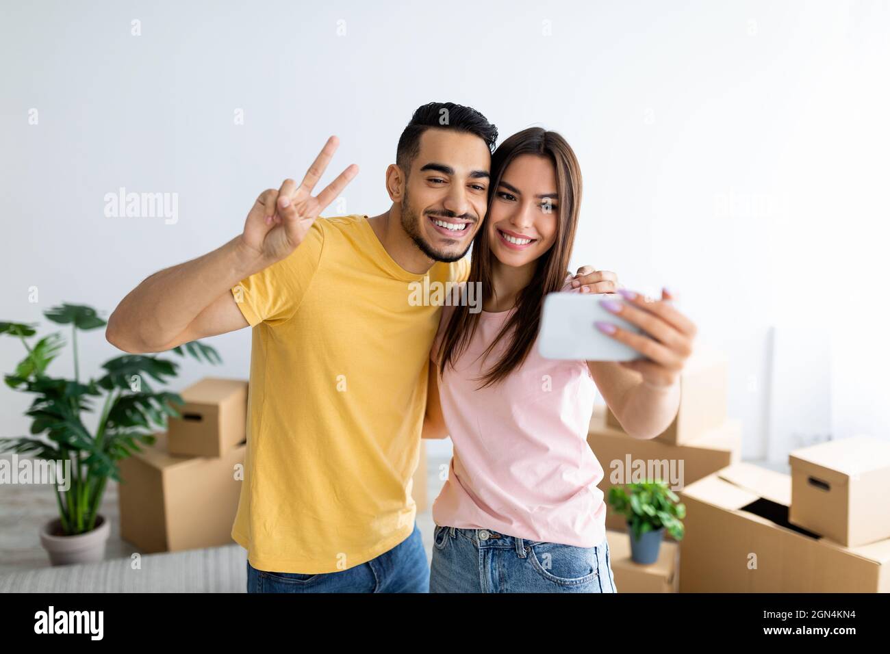 Sorridendo il ragazzo arabo e la sua ragazza caucasica che si trasferiscono a casa loro insieme, prendendo selfie tra scatole di cartone Foto Stock