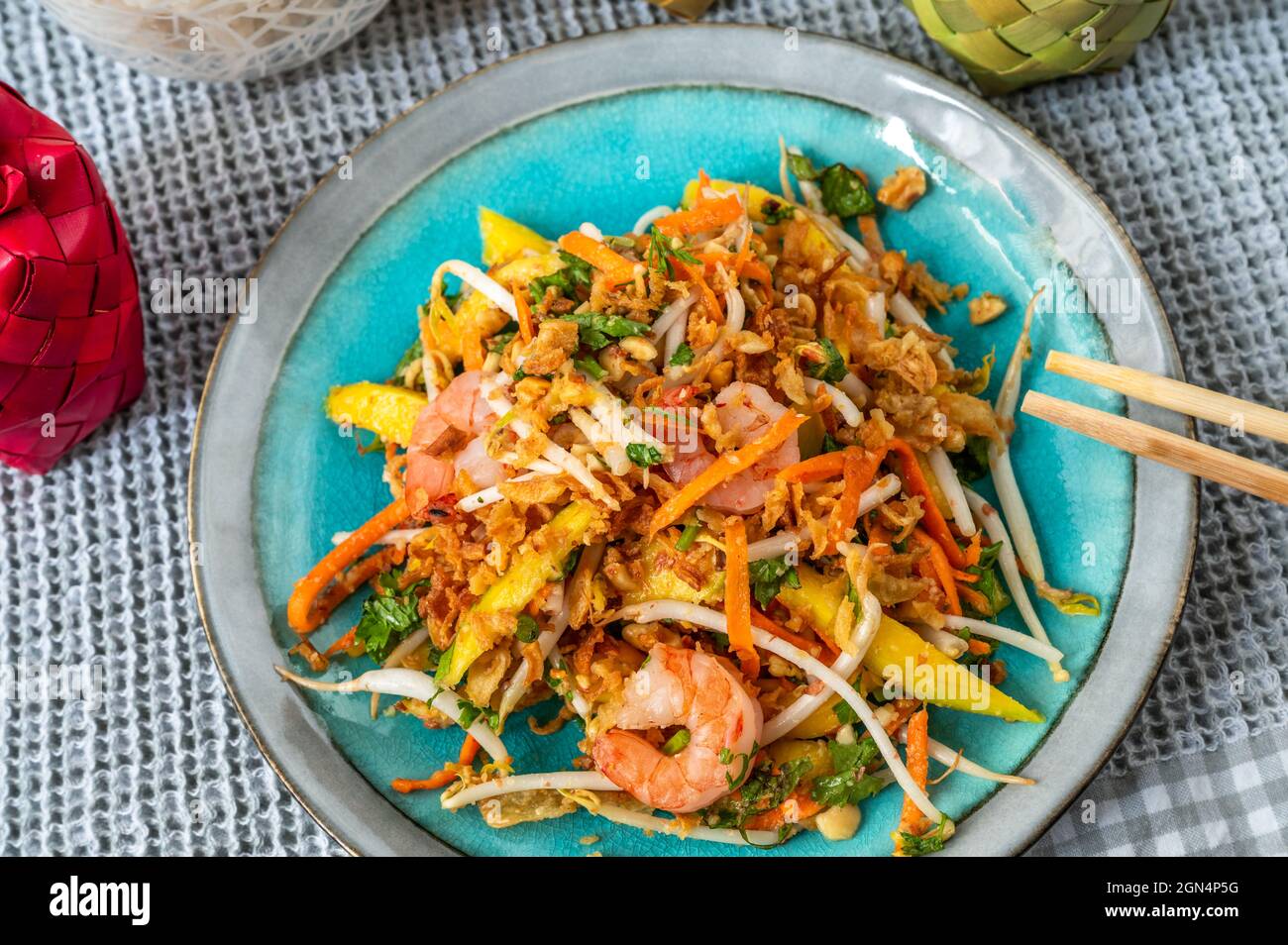 Insalata di mango dolce e aspro vietnamita con gamberi, cipolla fritta e germogli di verdure su piatto blu sul tavolo, bacchette. Foto Stock