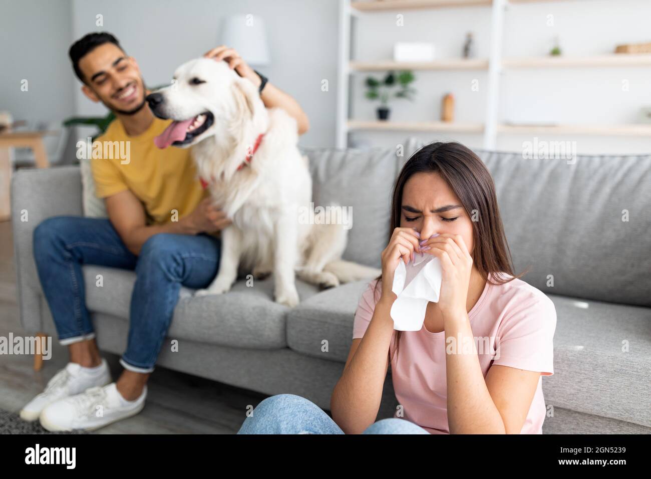 Signora caucasica malata che ha freddo, soffia il naso, starnutisce in carta tissue, il suo ragazzo arabo con cane da compagnia sullo sfondo Foto Stock