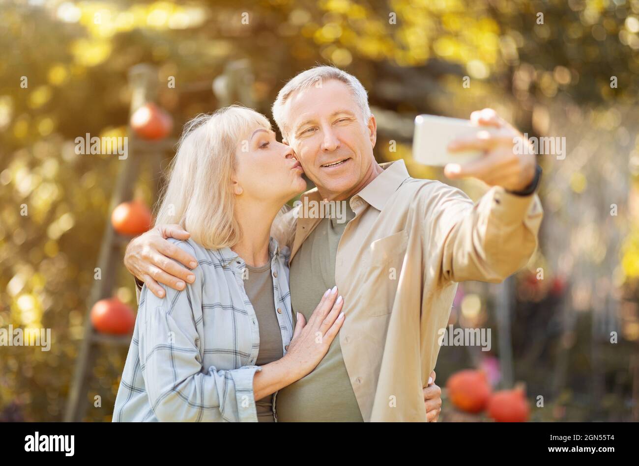 Coppia anziana che passa il tempo in giardino soleggiato insieme, prendendo selfie, donna baciando marito in guancia Foto Stock