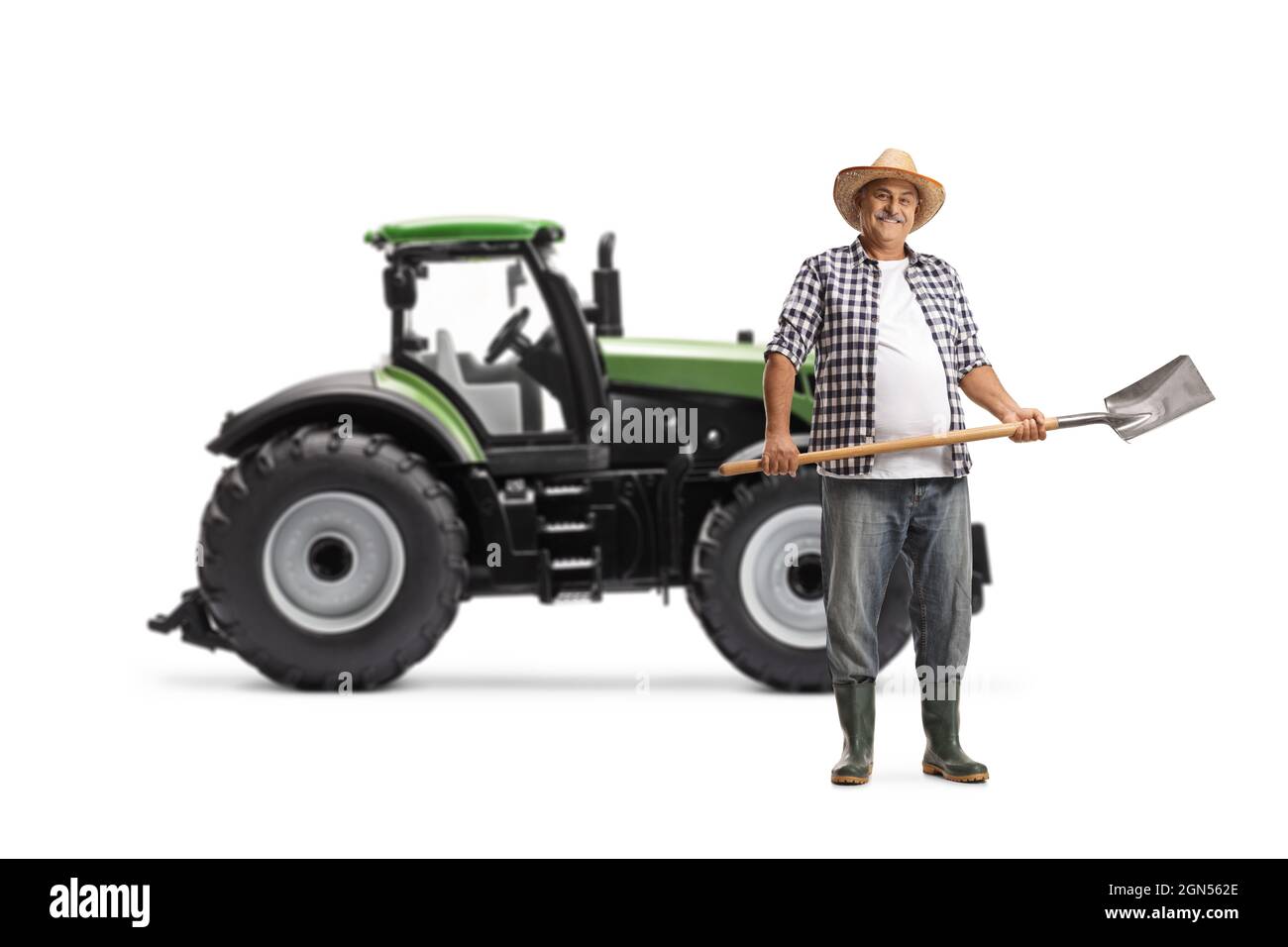 Ritratto a tutta lunghezza di un agricoltore maturo con una pala davanti ad un trattore verde isolato su sfondo bianco Foto Stock