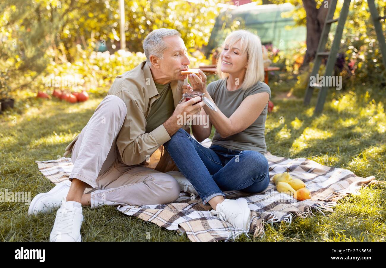 Coppia romantica anziana che ha pic-nic nel loro giardino, seduta su coperta e mangiare toast, trascorrere il tempo all'aperto Foto Stock