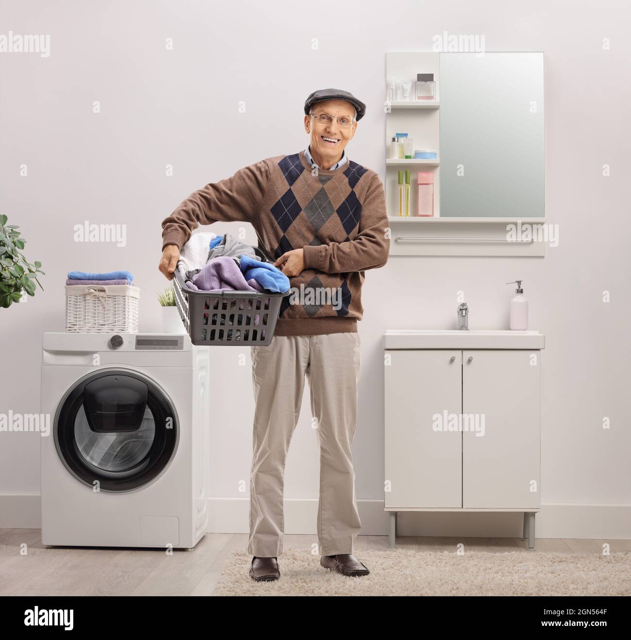 Uomo anziano sorridente che tiene un cesto di biancheria pieno di vestiti all'interno di un bagno Foto Stock