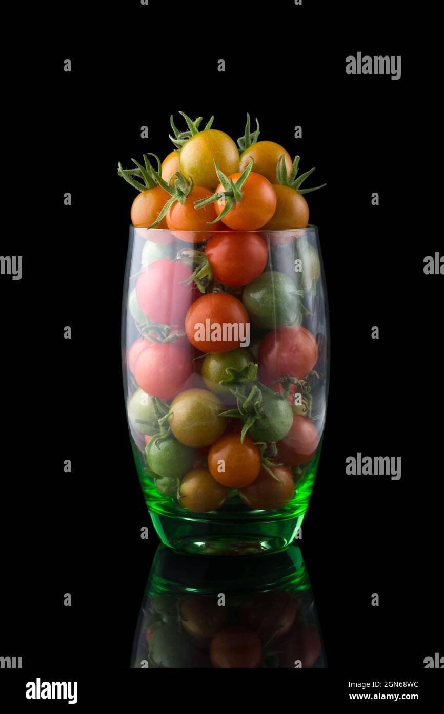 pomodori ciliegini, rossi, gialli e verdi piccoli pomodori rotondi in un bicchiere su una superficie riflettente, isolati su sfondo nero Foto Stock