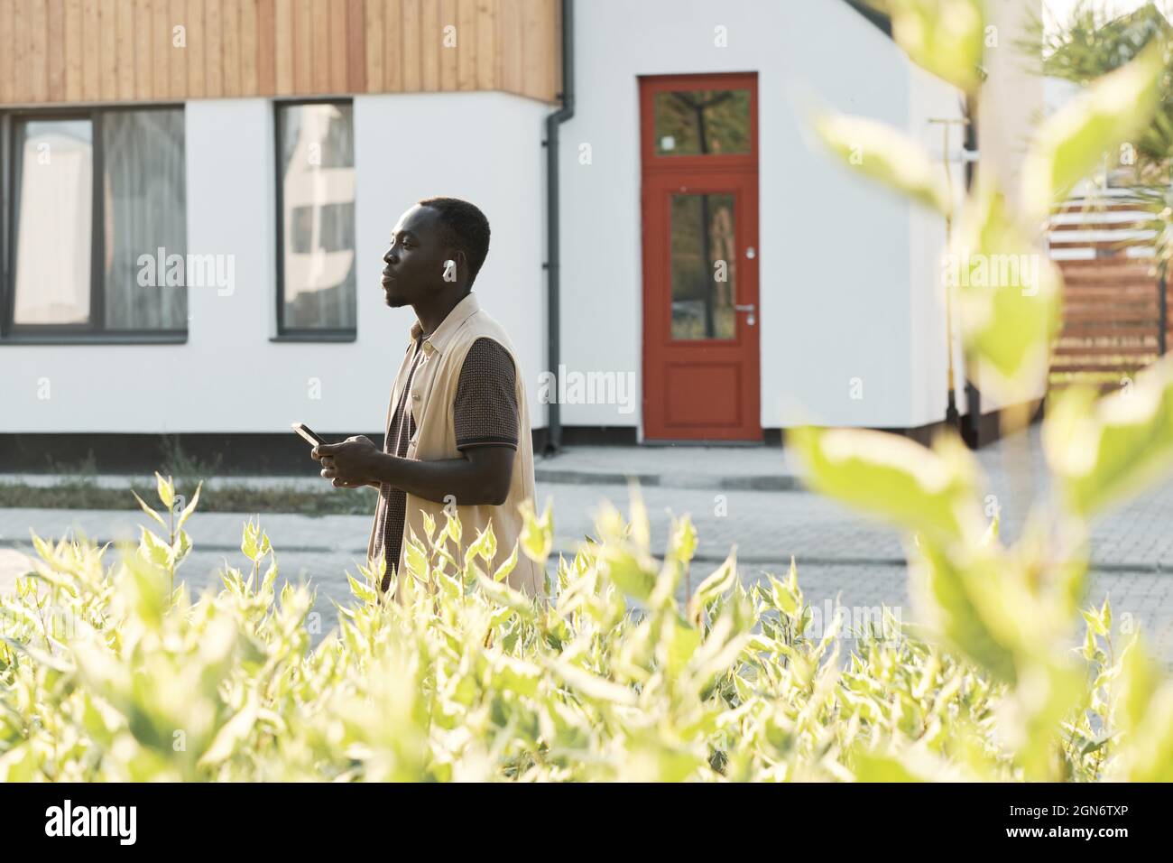 Scatto orizzontale laterale di un giovane afroamericano che cammina all'aperto con smartphone che ascolta musica utilizzando auricolari wireless Foto Stock