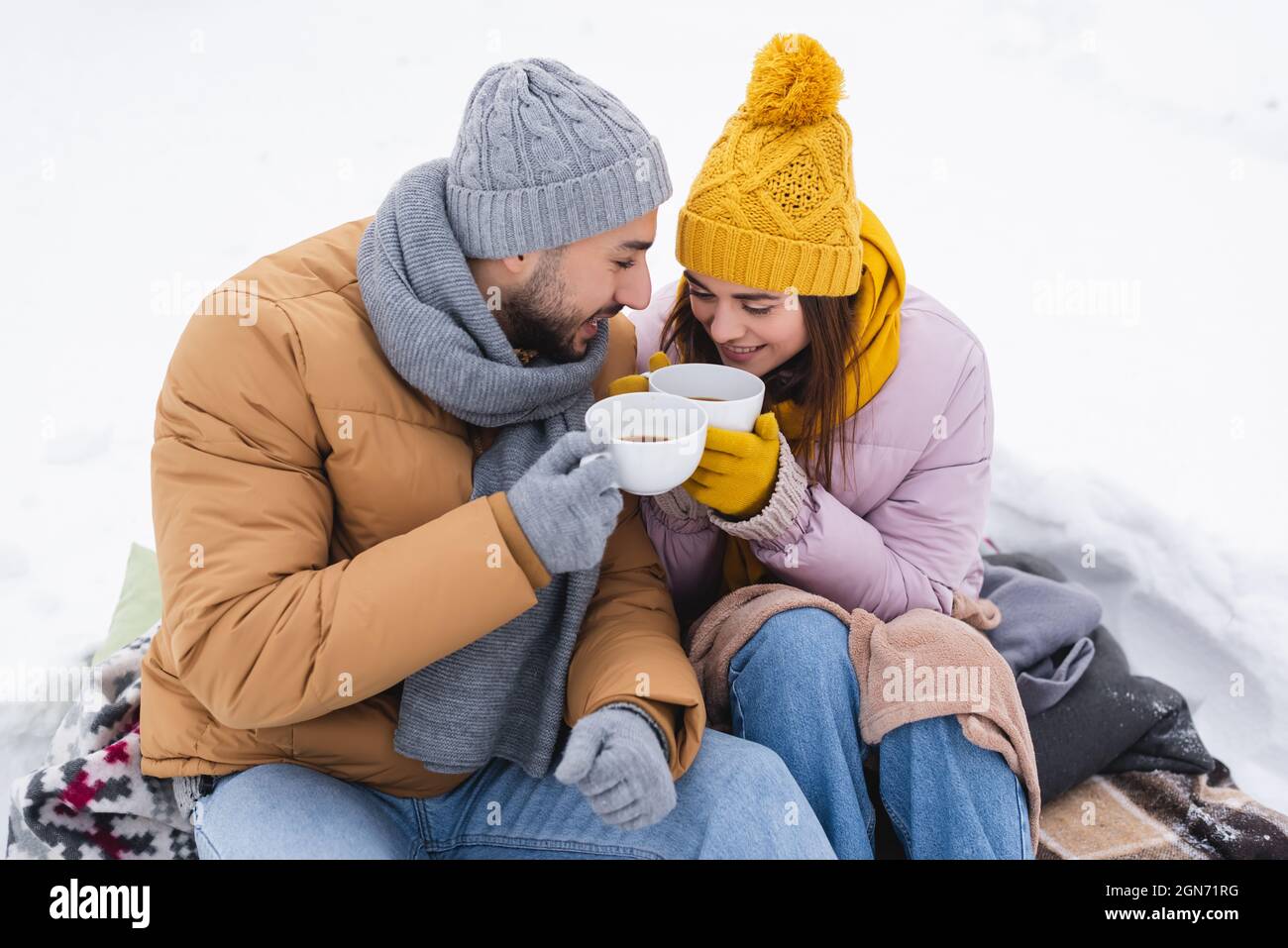 Coppia positiva che tiene tazze di caffè seduti su coperte sulla neve Foto Stock