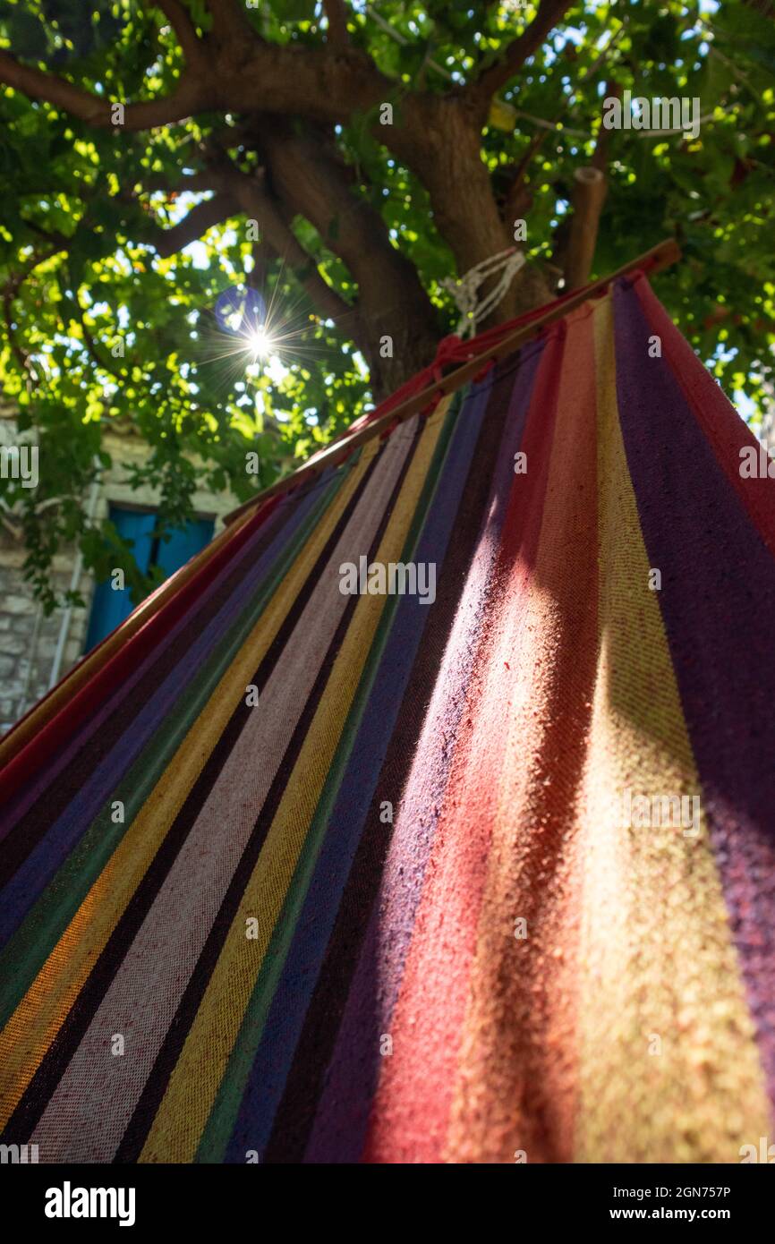 Dondolo in amaca colorata con sole che splende attraverso rami di alberi verdi, tempo di ricreazione estate. Vista verticale in cortile casa greco tradizionale con Foto Stock