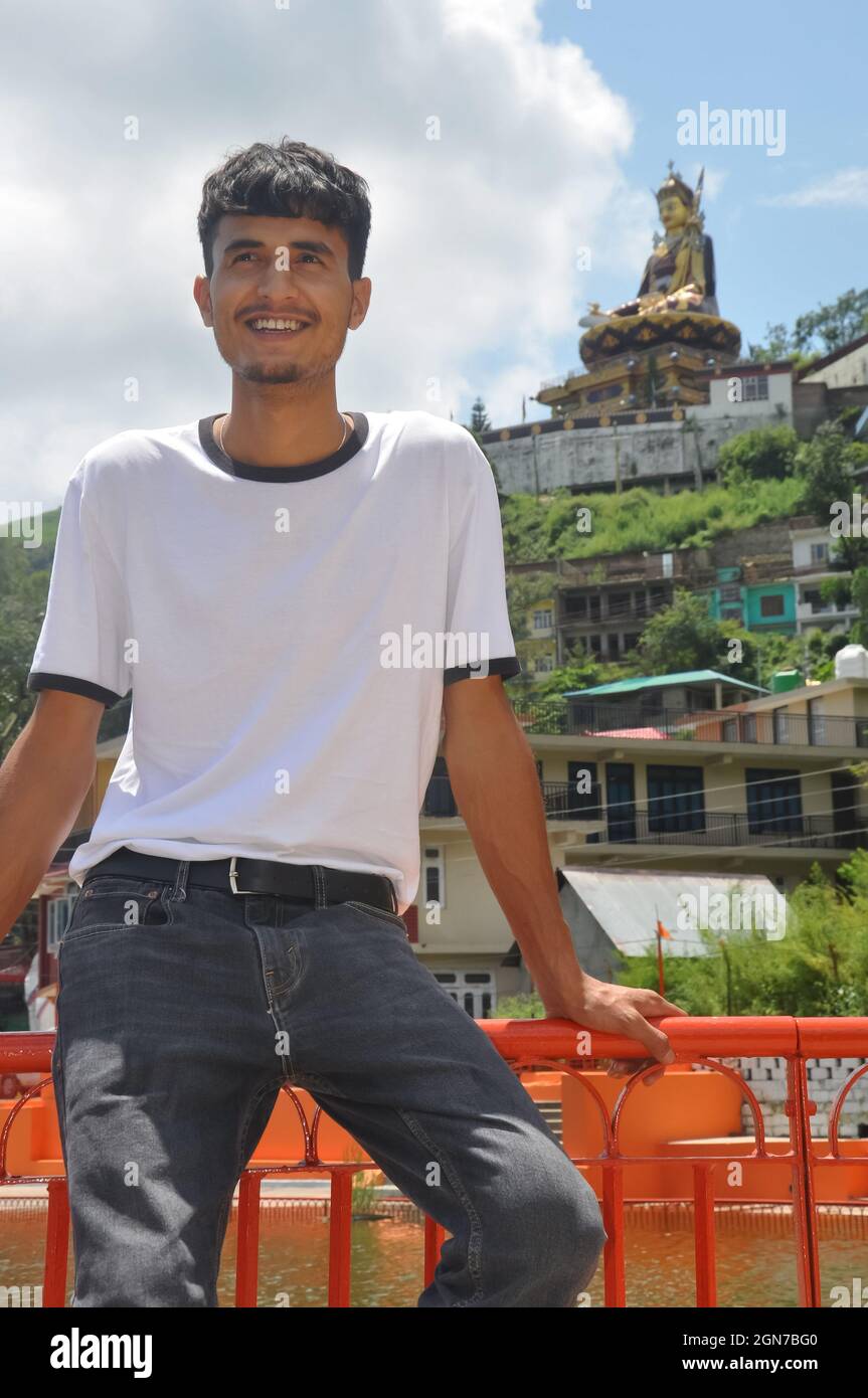 Un giovane sorridente seduto sulla barriera di sicurezza accanto al lago con lo sguardo lontano e bella zona collinare di Tso-Pema e la statua di Padmasambhava (Guru Rinpoche) in background Foto Stock