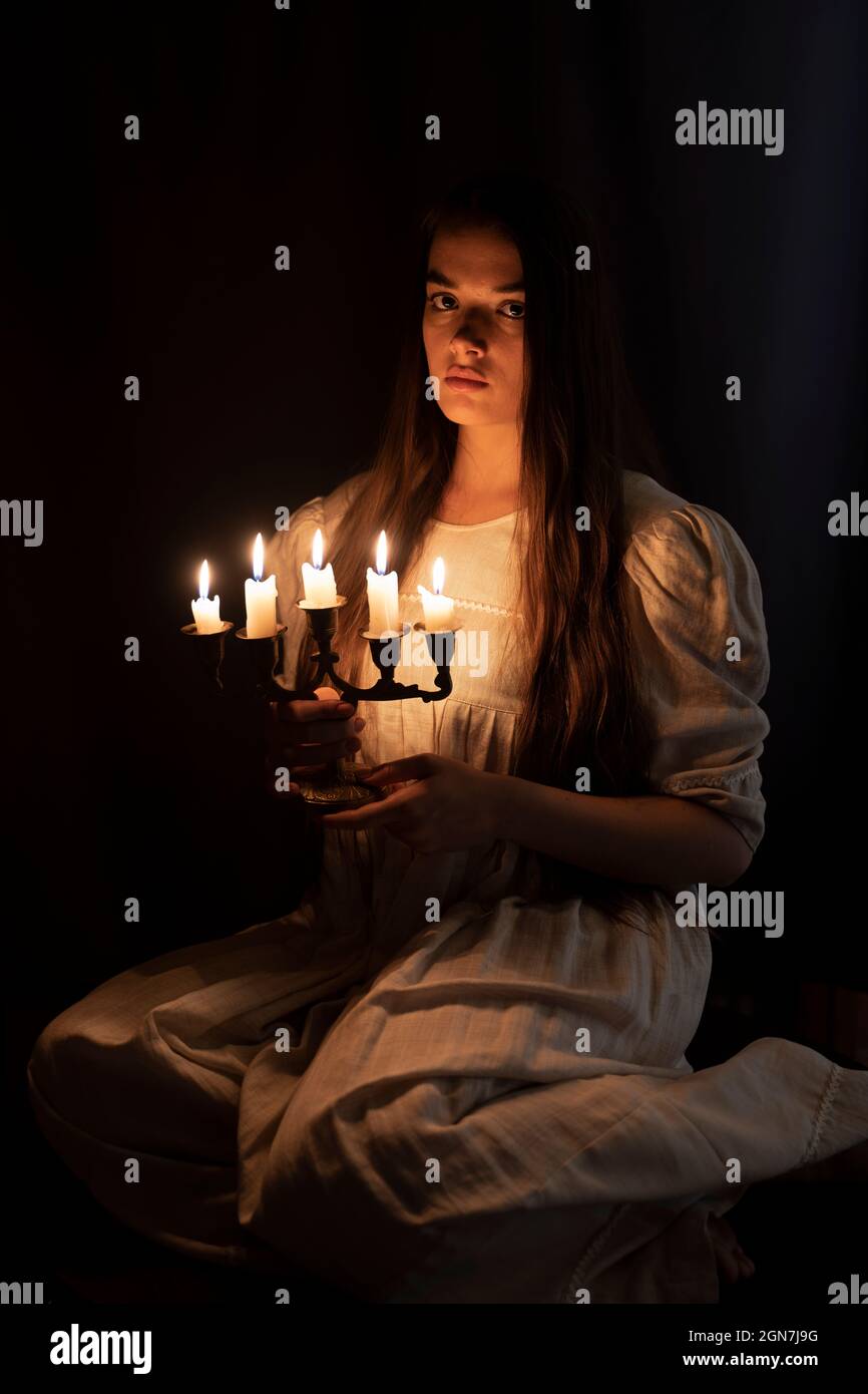 Una giovane ragazza in un vecchio vestito bianco seduto e tenendo una candela nelle sue mani. Sfondo scuro. Spaventoso concetto di orrore. Foto Stock