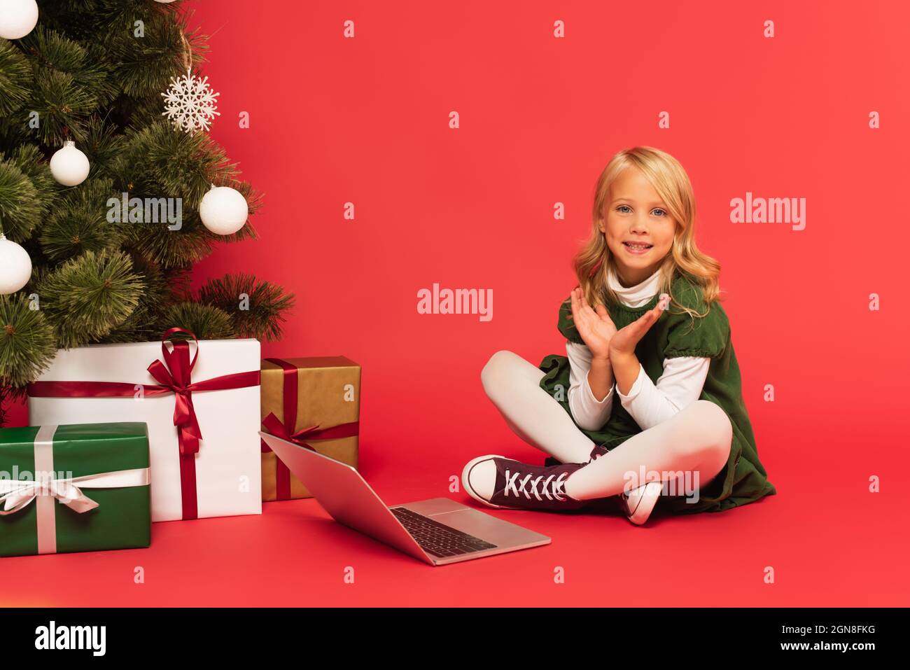 bambino felice che stringe le mani vicino al portatile e i regali sotto l'albero di natale su rosso Foto Stock