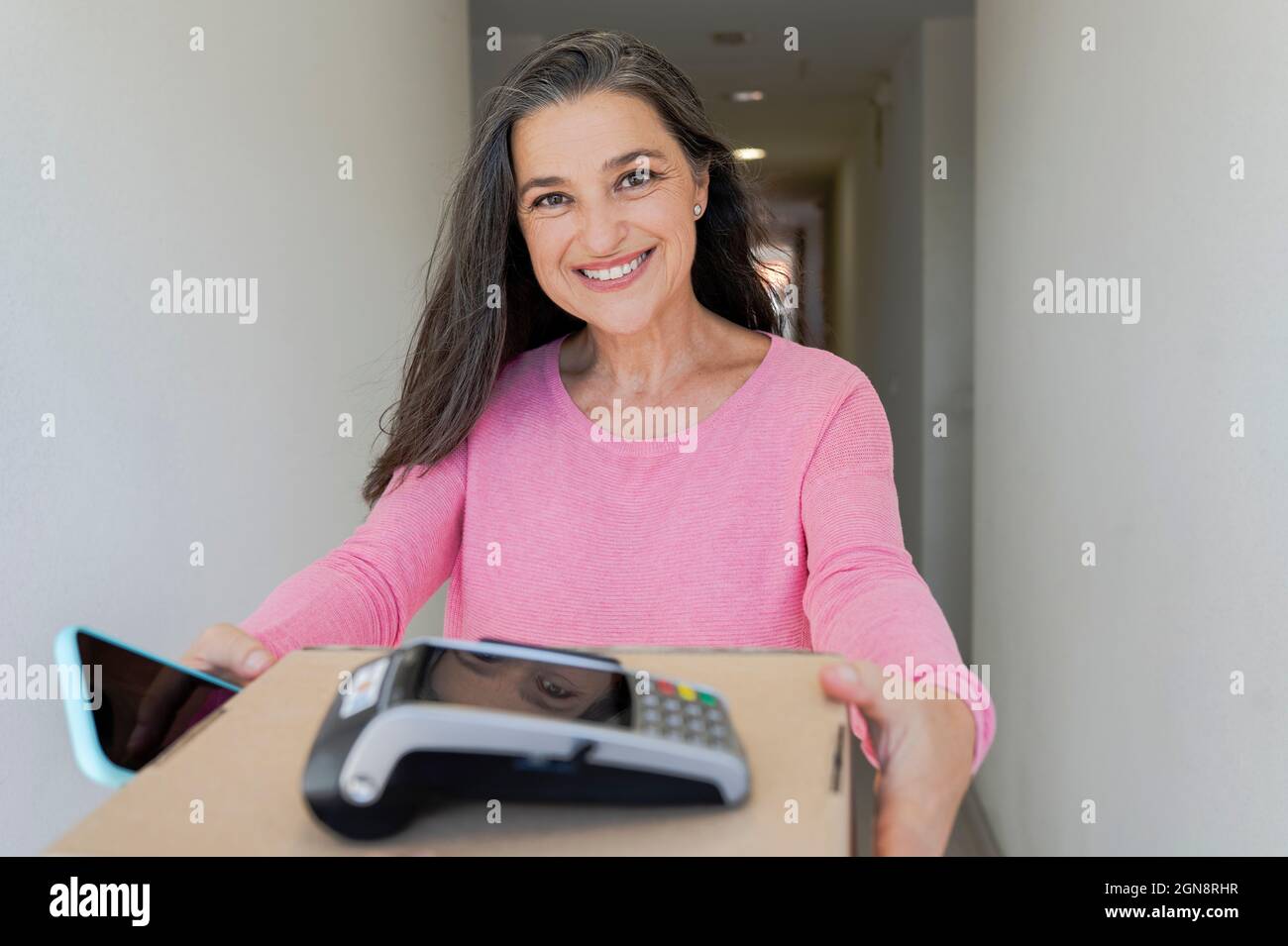 Donna sorridente con telefono cellulare che riceve il pacchetto in corridoio Foto Stock