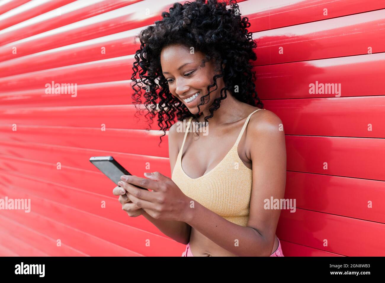 Happy Afro donna surf rete attraverso il telefono cellulare mentre si appoggia su otturatore rosso Foto Stock
