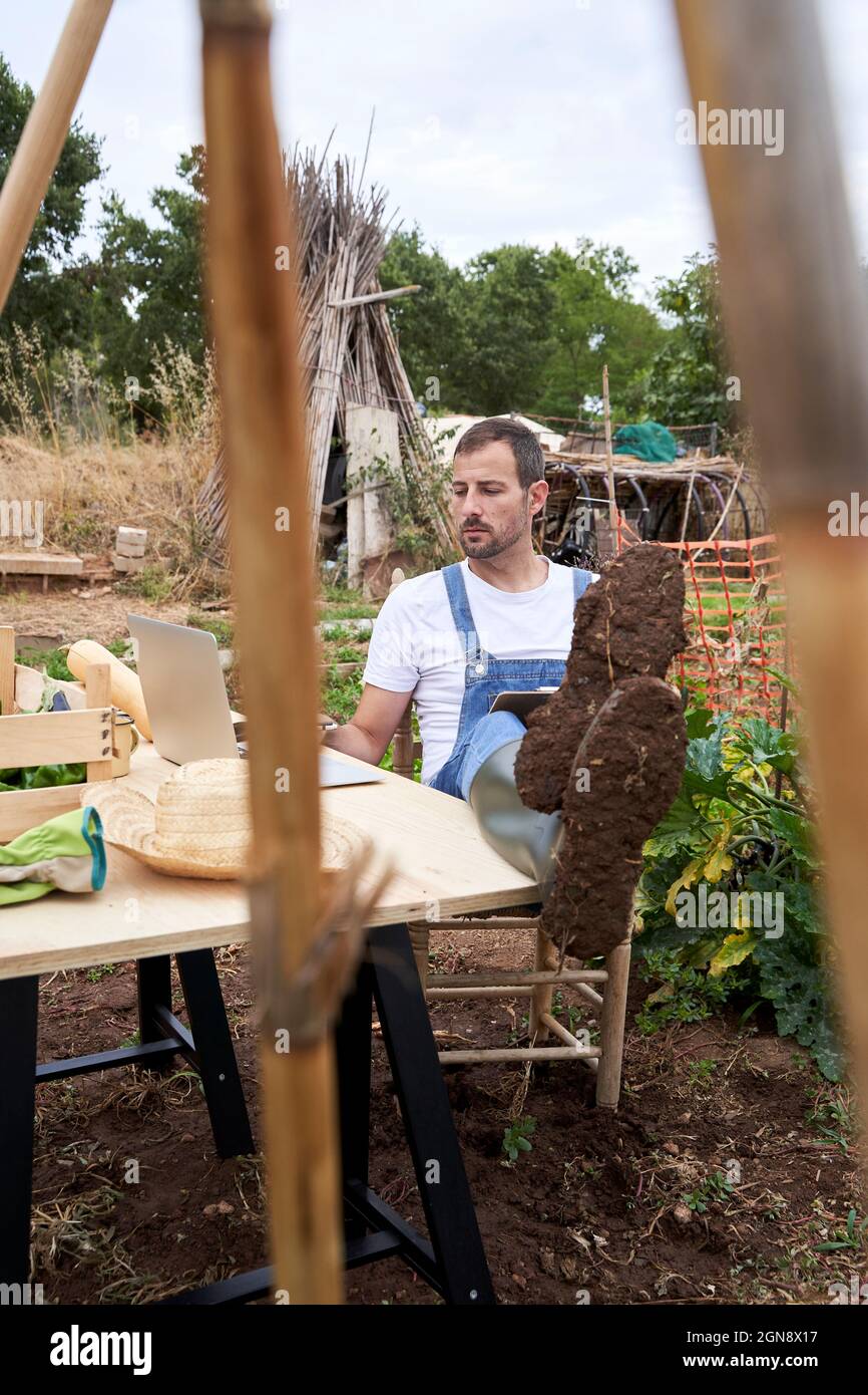 Agricoltore che utilizza il computer portatile mentre si siede con i piedi al tavolo in campo agricolo Foto Stock