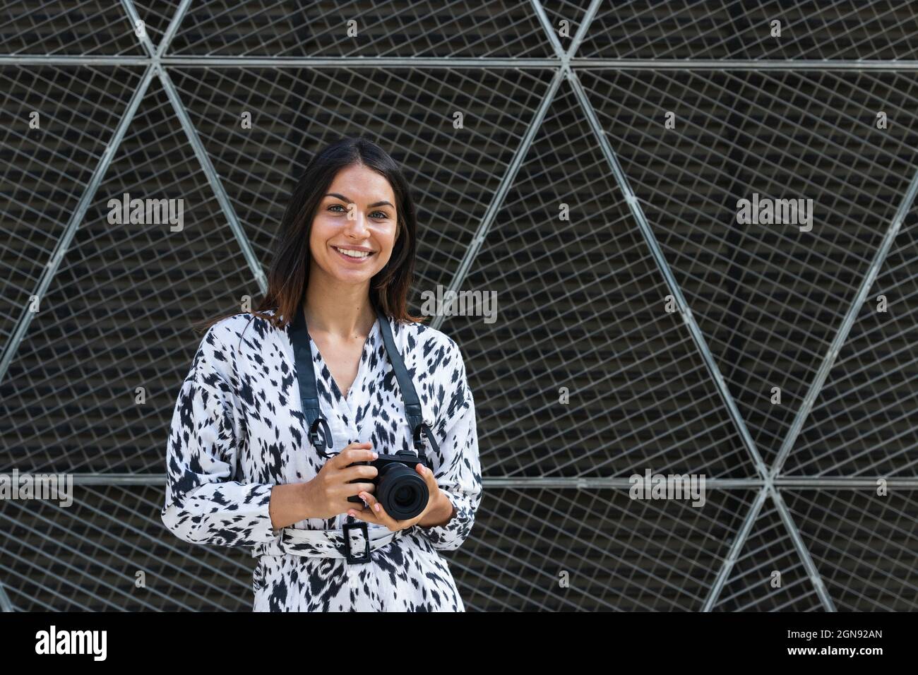 Sorridente giovane donna in piedi con la macchina fotografica di fronte alla recinzione Foto Stock