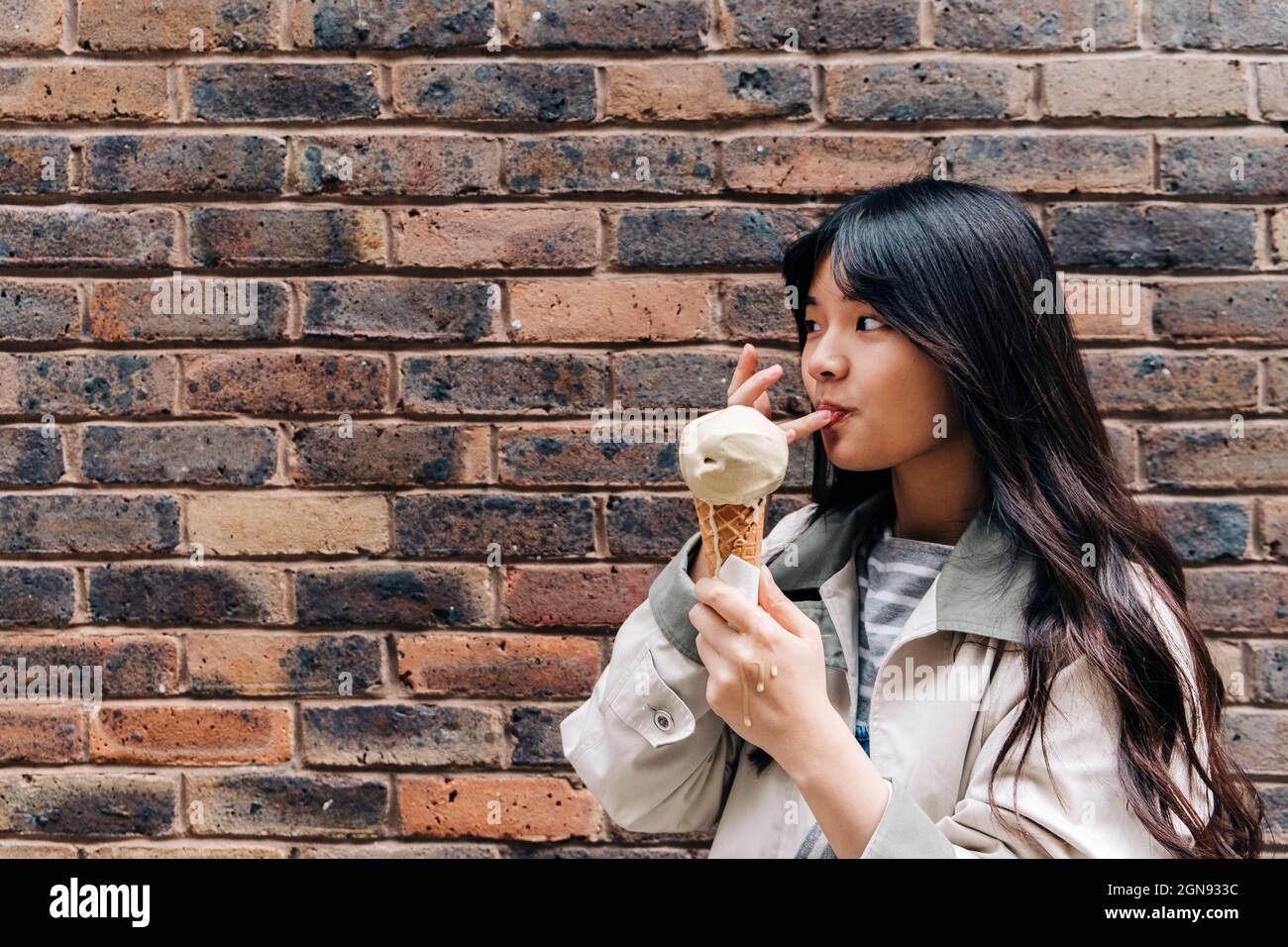 Donna che lecca il dito mentre tiene il gelato di fronte al muro di mattoni Foto Stock
