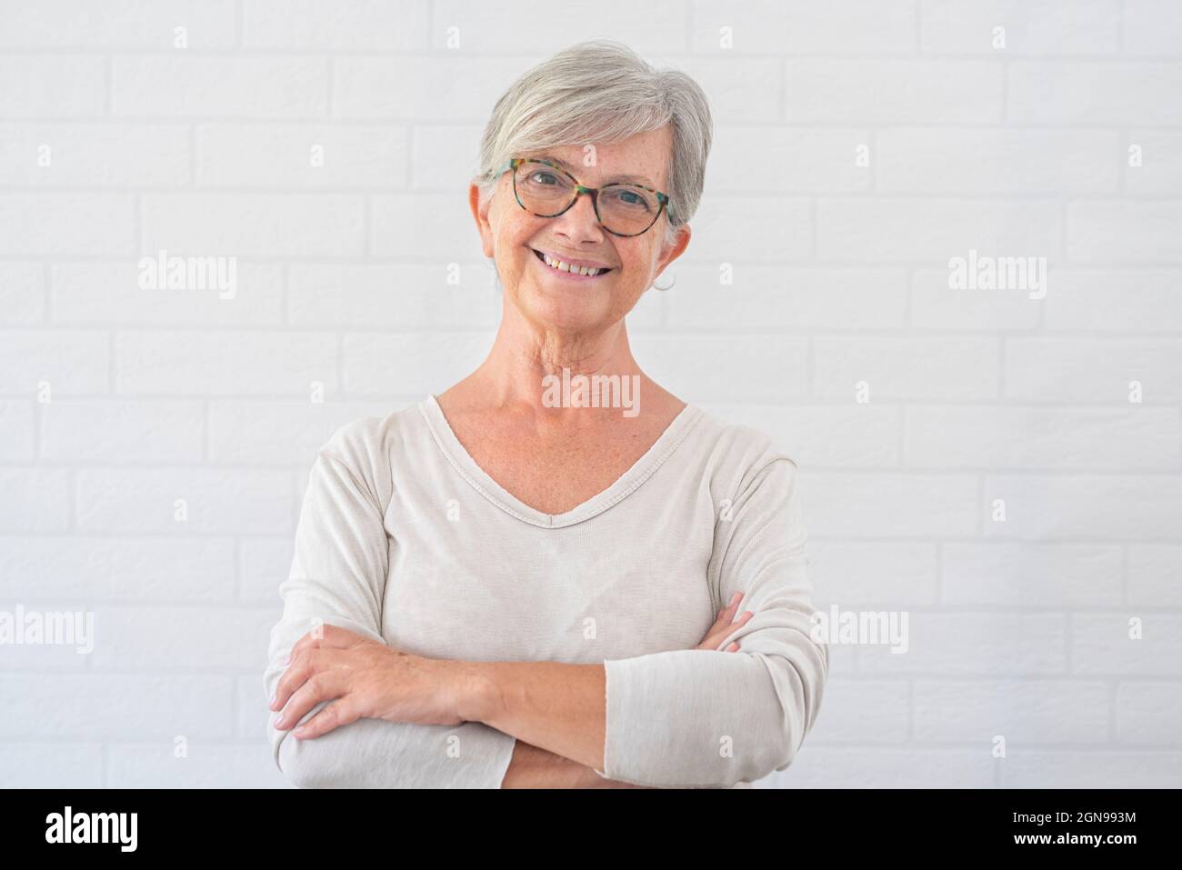 Donna anziana sorridente con le braccia incrociate di fronte al muro bianco Foto Stock