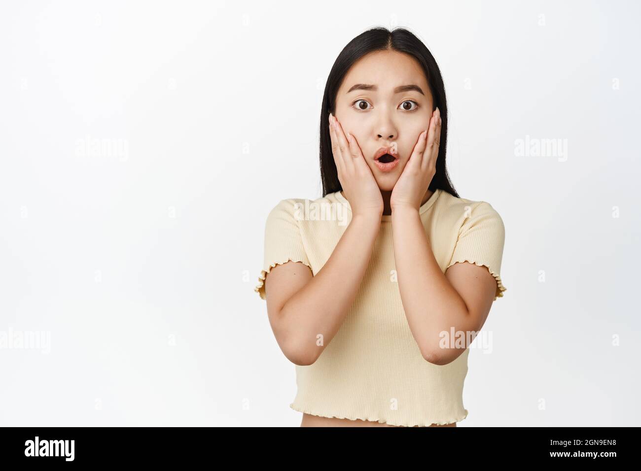 Scioccata e preoccupata ragazza asiatica che guarda preoccupato, fissando la fotocamera, tenendo le mani sul viso, in piedi su sfondo bianco Foto Stock