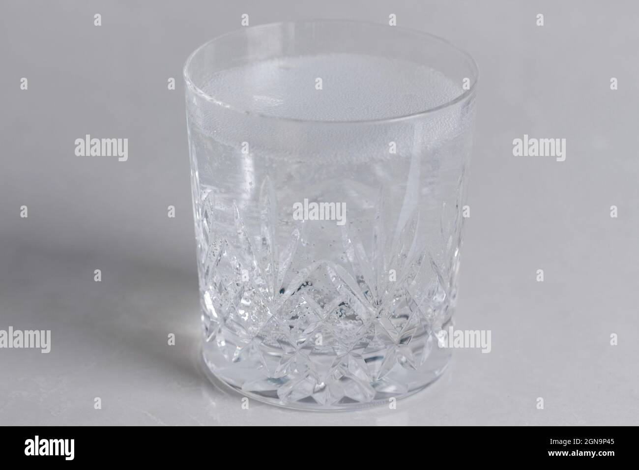 un bicchiere di cristallo finemente tagliato o vetro sparato riempito con acqua frizzante con uno strato di fizz o schiuma sopra su uno sfondo bianco con illuminazione naturale Foto Stock