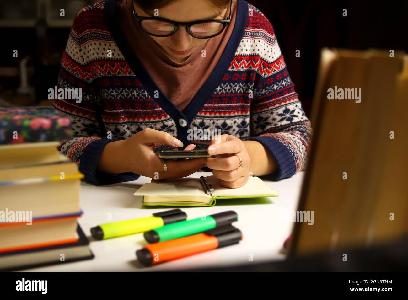 Giovane donna in occhiali studio di notte. Studente millenario che scatta foto al telefono. Preparazione dell'esame. Libri e marcatori sfocati in primo piano Foto Stock