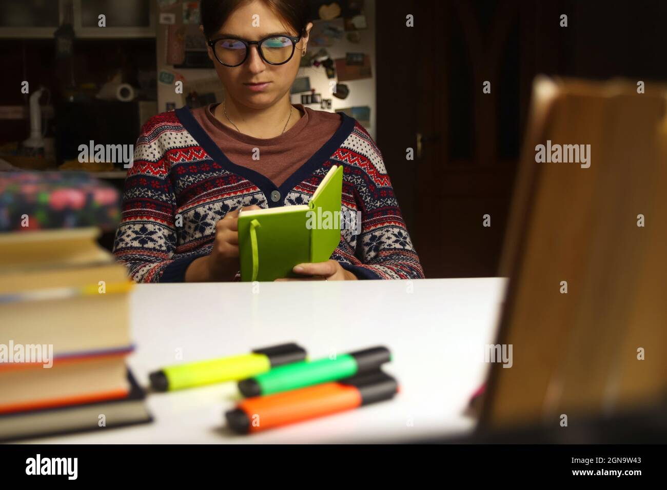 Giovane donna in occhiali studio di notte. Studente millenario che tiene e scrive in taccuino verde chiaro. Libri e marcatori sfocati in primo piano Foto Stock