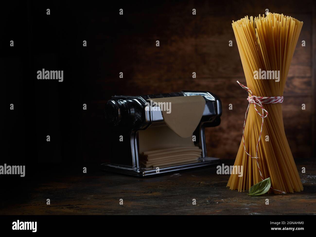 Mazzetto di spaghetti crudi su tavola in legno con macchina per pasta metallica e pasticceria fresca in studio scuro Foto Stock
