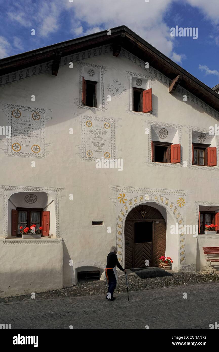 Un abitante anziano cammina con un bastone davanti ad una delle antiche case di Scuol Sot, la città vecchia costruita vicino al fiume alpino Inn o en a Scuol, Graubünden o Grigioni cantone, Svizzera. La casa restaurata ha due date: 1724 e 1986, l'anno in cui è stata ristrutturata. La facciata presenta fasce di decorazione sgraffito e un'iscrizione in Romansh, una lingua legacy dell'Impero Romano ancora parlato da circa la metà dei residenti di questa città termale della bassa Engadina Valley. Foto Stock