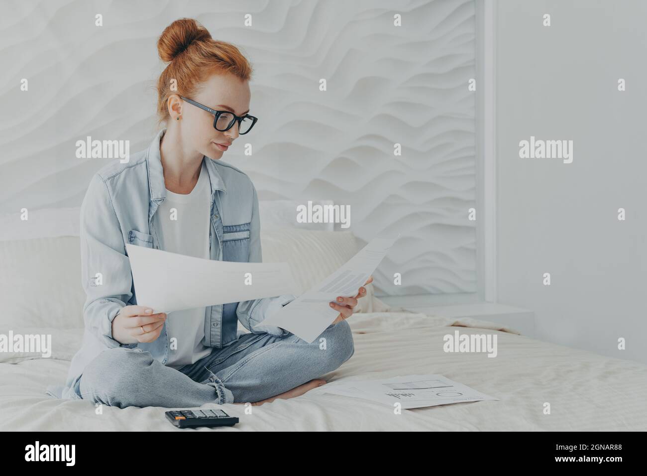 Giovane donna concentrata casalinga in occhiali che gestiscono il budget della famiglia mentre si siede a letto a casa Foto Stock