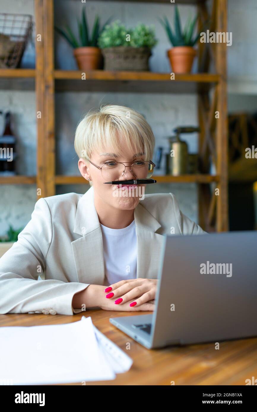 Foto di una giovane bionda con gli occhiali davanti a un computer portatile. È stanca di lavoro e di ingannare intorno durante una videochiamata in linea di affari. Lavorando da Foto Stock